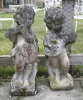 Lot 1062 - A pair of weathered composite stone figures of putti playing musical instruments